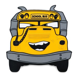 May include: A yellow school bus character patch with a grey bumper, grey exhaust pipes, and black tyres. The bus has a wide, grinning mouth with white teeth, narrow eyes, and a black chain detail draped across the bonnet. The text SCHOOL BUS is printed in black on the top yellow panel.