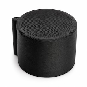 May include: A matte black cylindrical container with a textured surface featuring fine horizontal lines along the sides and a circular top lid with a distinct diagonal pattern. The object includes a small vertical tab on one side for opening.
