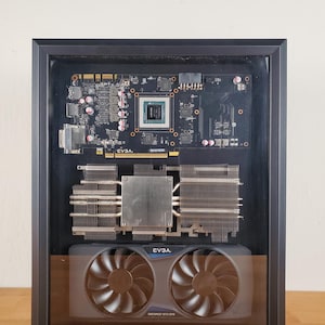 May include: A framed computer graphics card, featuring a black circuit board with silver heat sinks and two fans. The EVGA GeForce GTX 970 is visible. The frame is black and the background is a light wood.