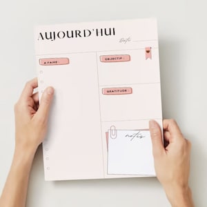 May include: A pink daily planner with sections for "A Faire", "Objectif", "Gratitude", and "Notes". The planner has a pink heart-shaped bookmark and a silver paper clip.