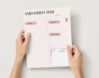 May include: A pink daily planner with sections for "A Faire", "Objectif", "Gratitude", and "Notes". The planner has a pink heart-shaped bookmark and a silver paper clip.