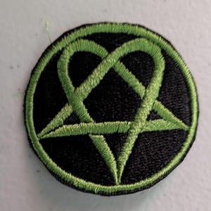 May include: Round black patch with a green heartagram design. The heartagram is a combination of a heart and a pentagram, with a green outline. The patch is likely an embroidered emblem, suitable for clothing or accessories.