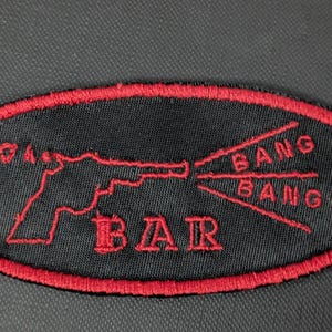 May include: Black oval patch with a red border. The patch features a red outline of a handgun with the words "BANG BANG" and "BAR" in red. The patch is likely for a jacket or bag.