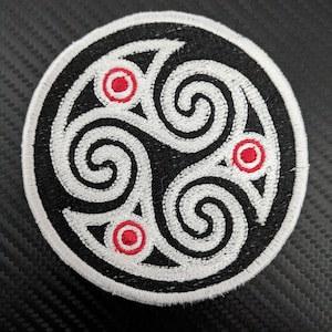 May include: Circular embroidered patch featuring a black background with a white Celtic knot design. The knot has three spirals and three red circles. The patch has a white border.