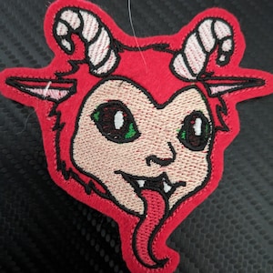 May include: Red embroidered patch of a cartoon goat with green eyes, a pink tongue sticking out, and white horns.