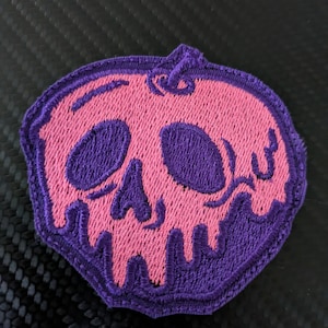 May include: A pink and purple embroidered patch shaped like an apple with a skull design. The skull has purple eyes and a purple outline.