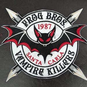 May include: Embroidered patch featuring a black bat with red accents, surrounded by text that reads "FROG BROS. 1987" and "VAMPIRE KILLERS SANTA CARLA." Two silver daggers are crossed behind the circular design.