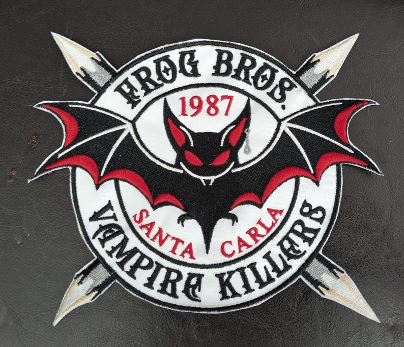 Puede incluir: Parche bordado con un murci&eacute;lago negro y detalles rojos, rodeado por el texto "FROG BROS. 1987" y "VAMPIRE KILLERS SANTA CARLA". Dos dagas plateadas se cruzan detr&aacute;s del dise&ntilde;o circular.