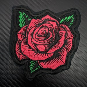 May include: A red embroidered rose patch with green leaves. The patch is on a black background.