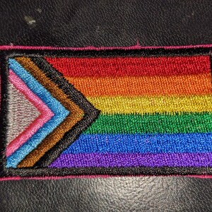 May include: A rainbow pride flag with a black, brown, light blue, pink, and white chevron in the corner. The flag is embroidered and has a pink border.
