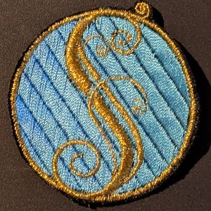 May include: A blue and gold embroidered patch with a stylized letter 'S' in the center. The letter is surrounded by a decorative border.