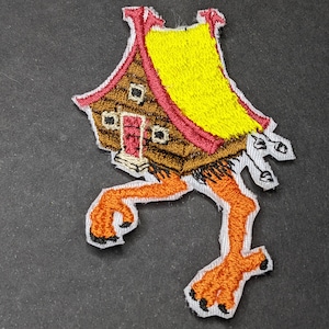 Puede incluir: Un parche bordado que representa una casa con piernas. La casa es marrón con un techo amarillo y una puerta roja. Las piernas son naranjas y tienen garras.