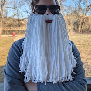 May include: A person wearing a white yarn beard and black sunglasses. The beard is long and covers the lower face. The person is wearing a grey sweatshirt and is standing outside.