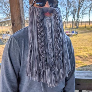 Peut inclure: Un accessoire de barbe et de moustache tressée grise et blanche. La barbe est faite de longs brins de fil, avec trois grandes tresses et une frange en bas. La personne porte des lunettes de soleil et un sweat-shirt gris.