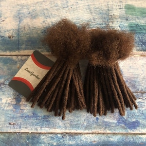 May include: Two bundles of brown synthetic dreadlocks, each with a fluffy end. The bundles are arranged side-by-side on a blue wooden surface. A small card with the text "LocsGarden" is visible in the lower left corner of the image.