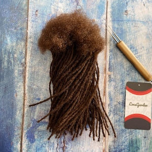 Puede incluir: Dreadlocks sintéticos marrones con una trama marrón, mostrados sobre un fondo de madera azul. También se muestra una herramienta de madera con una aguja de metal, junto con una etiqueta blanca con el texto "LocsGarden".