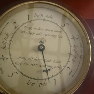 May include: A round tide clock with a brass-colored metal frame and a cream-colored face. The dial features black text, including "The Nature Company" logo, and markings for high tide, low tide, and half tide, with a single black indicator hand.
