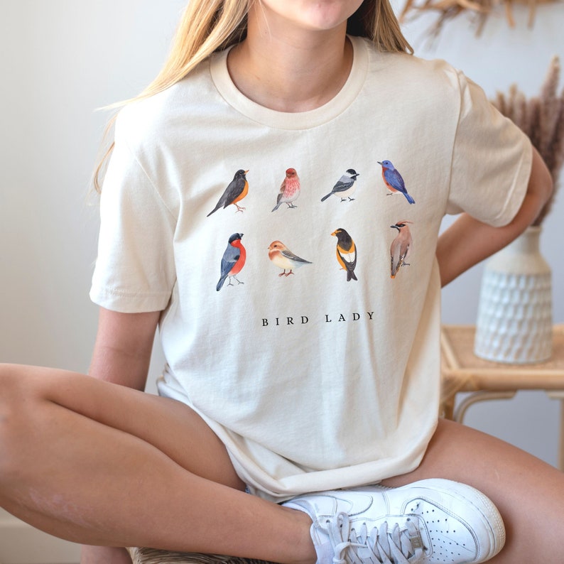 May include: A white t-shirt with a graphic of nine different birds in watercolor style. The text "BIRD LADY" is printed below the birds.