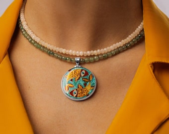 Può includere: Una donna indossa una giacca gialla con una collana a strati che presenta una fila di perline verdi e bianche e un ciondolo d'argento con un design di pesce turchese e oro.