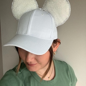 May include: A light blue baseball cap with attached mouse ears. The ears are round, padded, and feature a floral pattern in light gray and white, trimmed with a silver rope. The cap is a solid light blue color.