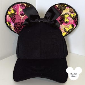 Puede incluir: Gorra de b&eacute;isbol negra con orejas de rat&oacute;n adjuntas. Las orejas presentan un estampado de guepardo rosa, amarillo y negro con detalles de lazo. Las orejas est&aacute;n ribeteadas con cuerda negra y un lazo negro est&aacute; centrado en la gorra. El texto "Cheetah Mickey" est&aacute; en la esquina inferior derecha.