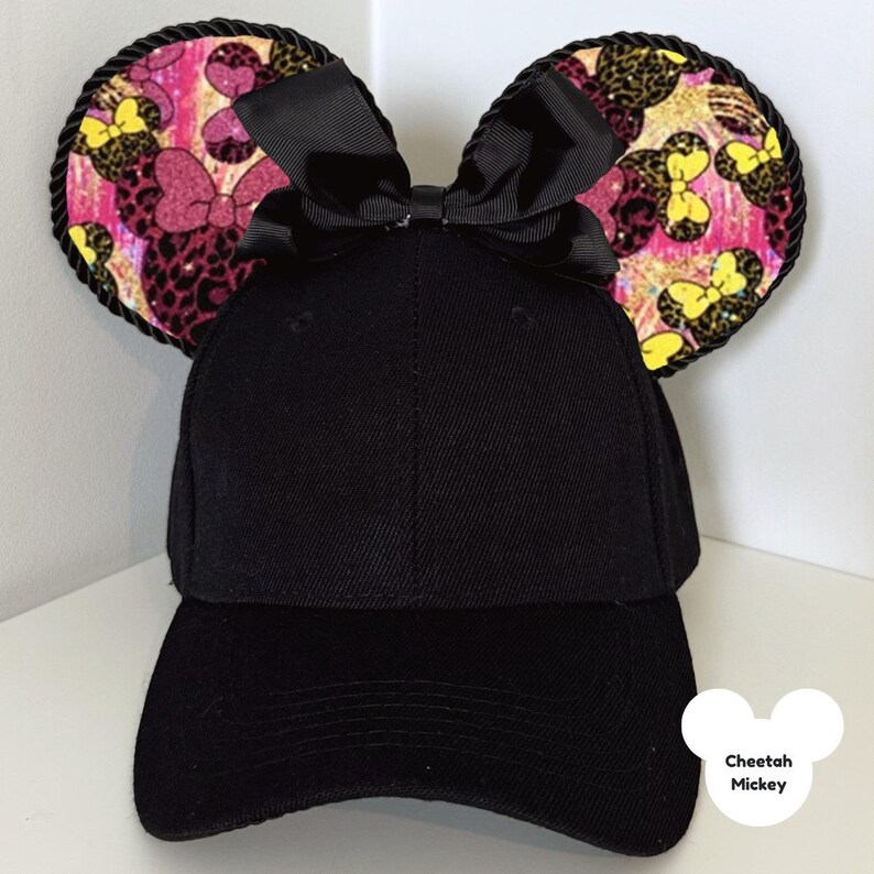 Puede incluir: Gorra de b&eacute;isbol negra con orejas de rat&oacute;n adjuntas. Las orejas presentan un estampado de guepardo rosa, amarillo y negro con detalles de lazo. Las orejas est&aacute;n ribeteadas con cuerda negra y un lazo negro est&aacute; centrado en la gorra. El texto "Cheetah Mickey" est&aacute; en la esquina inferior derecha.