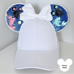 Puede incluir: Gorra de b&eacute;isbol blanca con orejas decorativas con un tema espacial azul, que incluye estrellas y criaturas marinas. Un gran lazo blanco se encuentra en la parte superior de la gorra. El texto "Whimsy Mickey" est&aacute; en la parte inferior derecha.