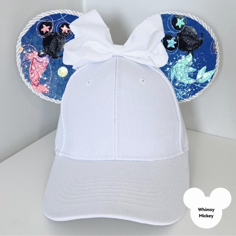 Puede incluir: Gorra de b&eacute;isbol blanca con orejas decorativas con un tema espacial azul, que incluye estrellas y criaturas marinas. Un gran lazo blanco se encuentra en la parte superior de la gorra. El texto "Whimsy Mickey" est&aacute; en la parte inferior derecha.