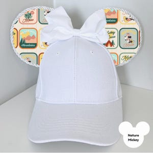 Puede incluir: Gorra de b&eacute;isbol blanca con orejas de rat&oacute;n decorativas. Las orejas presentan un patr&oacute;n colorido con texto e ilustraciones de un rat&oacute;n, monta&ntilde;as y escenas de acampada. Un lazo blanco se encuentra en la parte superior de la gorra. El texto "Nature Mickey" es visible.