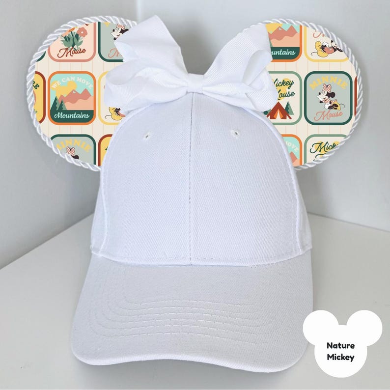 Puede incluir: Gorra de b&eacute;isbol blanca con orejas de rat&oacute;n decorativas. Las orejas presentan un patr&oacute;n colorido con texto e ilustraciones de un rat&oacute;n, monta&ntilde;as y escenas de acampada. Un lazo blanco se encuentra en la parte superior de la gorra. El texto "Nature Mickey" es visible.