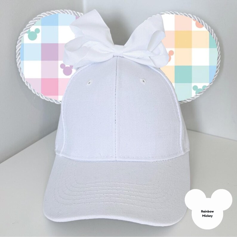 Puede incluir: Una gorra de b&eacute;isbol blanca con orejas de rat&oacute;n adjuntas. Las orejas son redondas con un patr&oacute;n de cuadros pastel y un ribete de cuerda blanca. Un gran lazo blanco se encuentra en la parte superior de la gorra. El texto "Rainbow Mickey" est&aacute; en la parte inferior derecha.