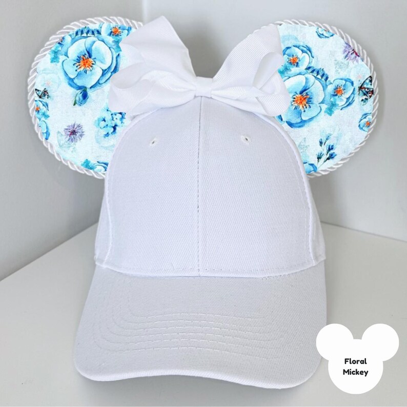 Puede incluir: Gorra de b&eacute;isbol blanca con orejas de Mickey Mouse florales. Las orejas presentan un estampado floral azul claro con mariposas y un ribete de cuerda blanca. Un gran lazo blanco se asienta sobre la gorra. El texto "Floral Mickey" est&aacute; en la parte inferior derecha.