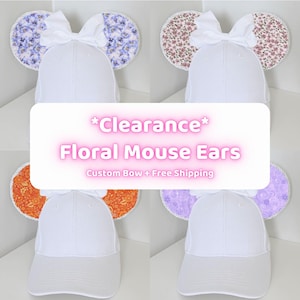 Op de afbeelding: Witte baseballpetten met bloemen muizenoren in verschillende patronen, waaronder blauw, roze, oranje en paars. Elke pet heeft een witte strik. De afbeelding bevat de tekst "*Clearance* Floral Mouse Ears".