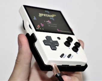 May include: A white and black handheld video game console with a black and gray directional pad, four black buttons, and a screen displaying a pixelated game. The console has a headphone jack on the bottom right side.