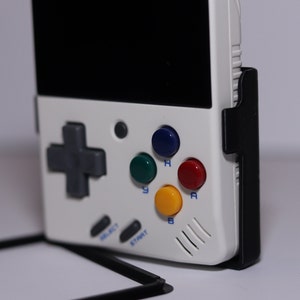 K&ouml;nnte beinhalten: Eine wei&szlig;e Handheld-Videospielkonsole mit einem schwarzen Bildschirm, einem grauen Steuerkreuz und vier farbigen Kn&ouml;pfen: gr&uuml;n, blau, rot und gelb. Die Konsole hat einen schwarzen St&auml;nder an der Seite.