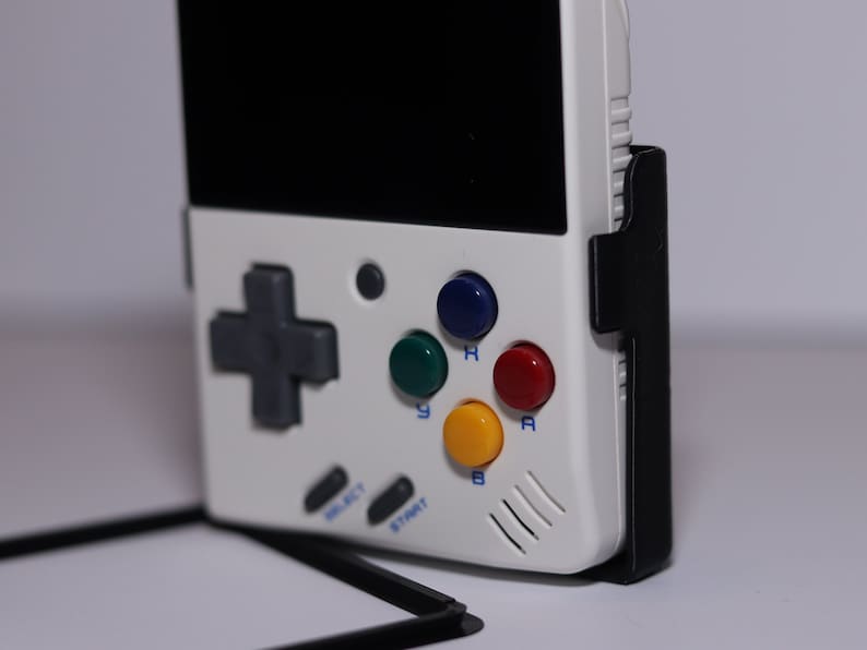 K&ouml;nnte beinhalten: Eine wei&szlig;e Handheld-Videospielkonsole mit einem schwarzen Bildschirm, einem grauen Steuerkreuz und vier farbigen Kn&ouml;pfen: gr&uuml;n, blau, rot und gelb. Die Konsole hat einen schwarzen St&auml;nder an der Seite.