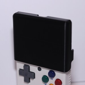 K&ouml;nnte beinhalten: Eine wei&szlig;e Handheld-Spielkonsole mit einem schwarzen Bildschirm, einem grauen Steuerkreuz und vier farbigen Kn&ouml;pfen. Die Kn&ouml;pfe sind gr&uuml;n, blau, rot und gelb.