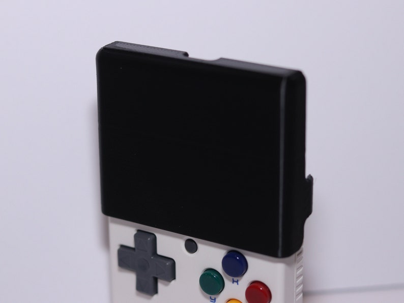 K&ouml;nnte beinhalten: Eine wei&szlig;e Handheld-Spielkonsole mit einem schwarzen Bildschirm, einem grauen Steuerkreuz und vier farbigen Kn&ouml;pfen. Die Kn&ouml;pfe sind gr&uuml;n, blau, rot und gelb.