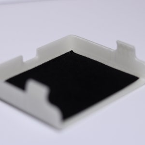 K&ouml;nnte beinhalten: Ein wei&szlig;es Plastikgeh&auml;use mit schwarzem Filzbezug. Das Geh&auml;use ist f&uuml;r die Aufbewahrung eines kleinen Gegenstands wie eines Telefons oder einer Uhr konzipiert.