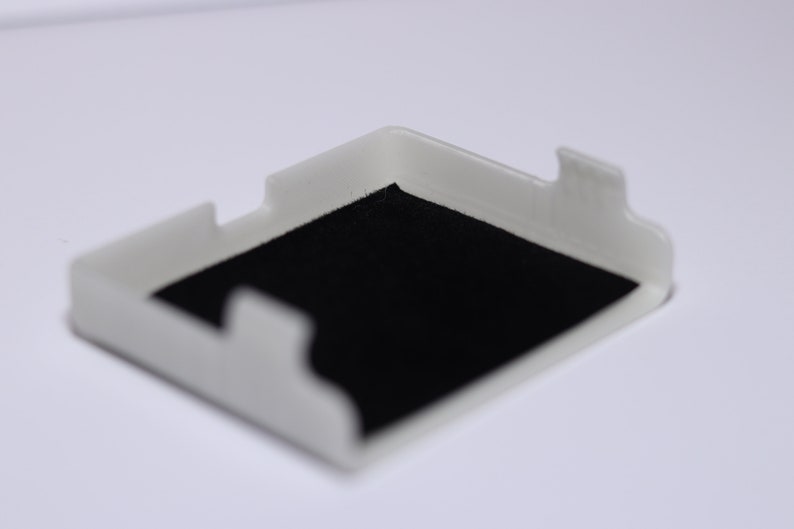 K&ouml;nnte beinhalten: Ein wei&szlig;es Plastikgeh&auml;use mit schwarzem Filzbezug. Das Geh&auml;use ist f&uuml;r die Aufbewahrung eines kleinen Gegenstands wie eines Telefons oder einer Uhr konzipiert.