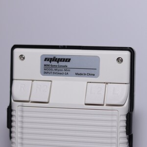 K&ouml;nnte beinhalten: Eine wei&szlig;e Handheld-Spielkonsole mit schwarzem Rahmen und vier Kn&ouml;pfen. Die Konsole hat ein silbernes Etikett mit der Aufschrift "miyoo MINI Game Console MODEL:Miyoo-Mini INPUT:5V(max)-1A Made In China".