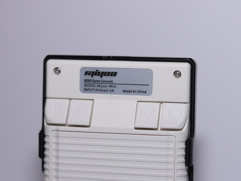 K&ouml;nnte beinhalten: Eine wei&szlig;e Handheld-Spielkonsole mit schwarzem Rahmen und vier Kn&ouml;pfen. Die Konsole hat ein silbernes Etikett mit der Aufschrift "miyoo MINI Game Console MODEL:Miyoo-Mini INPUT:5V(max)-1A Made In China".