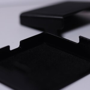 K&ouml;nnte beinhalten: Eine schwarze rechteckige Box mit einem Deckel, der sich &ouml;ffnet und einen schwarzen, samtigen Innenraum enth&uuml;llt. Die Box ist aus einem harten, strapazierf&auml;higen Material gefertigt und hat ein elegantes, modernes Design.