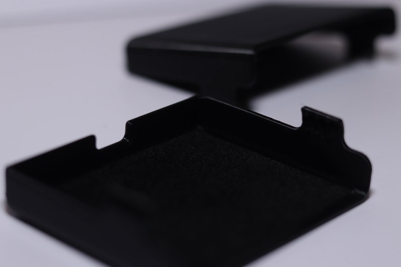 K&ouml;nnte beinhalten: Eine schwarze rechteckige Box mit einem Deckel, der sich &ouml;ffnet und einen schwarzen, samtigen Innenraum enth&uuml;llt. Die Box ist aus einem harten, strapazierf&auml;higen Material gefertigt und hat ein elegantes, modernes Design.