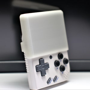 K&ouml;nnte beinhalten: Eine wei&szlig;e Handheld-Videospielkonsole mit einem grauen Steuerkreuz und vier schwarzen Kn&ouml;pfen. Die Konsole hat einen wei&szlig;en Bildschirm und blauen Text, der "SELECT" und "START" lautet.