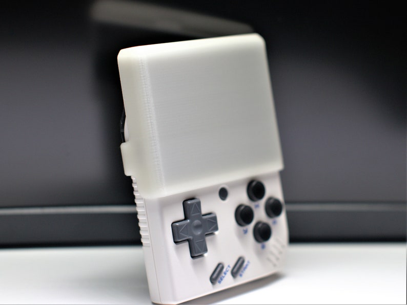 K&ouml;nnte beinhalten: Eine wei&szlig;e Handheld-Videospielkonsole mit einem grauen Steuerkreuz und vier schwarzen Kn&ouml;pfen. Die Konsole hat einen wei&szlig;en Bildschirm und blauen Text, der "SELECT" und "START" lautet.
