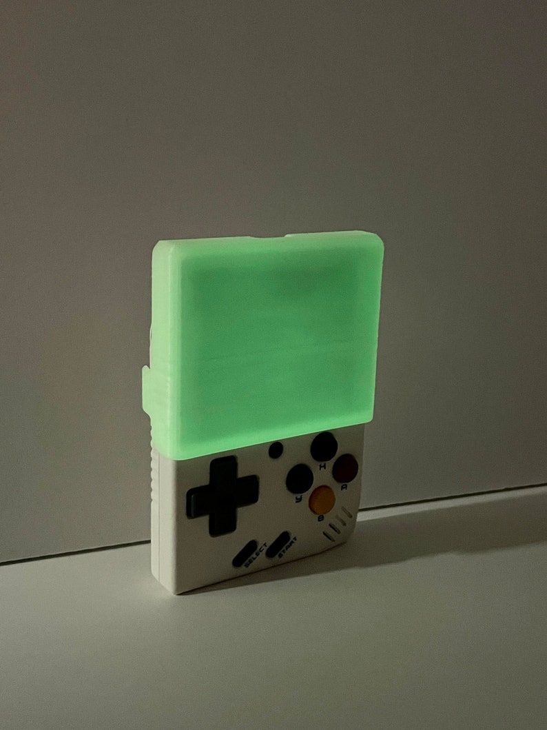 K&ouml;nnte beinhalten: Eine wei&szlig;e Handheld-Spielkonsole mit einem gr&uuml;n leuchtenden Bildschirm. Die Konsole hat ein Steuerkreuz, vier Tasten und einen Start- und einen Select-Knopf.