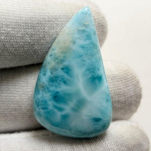 Könnte beinhalten: Ein hellblauer, perlmuttartiger Cabochon-Edelstein mit einem einzigartigen, wirbelnden Muster. Der Stein hat eine glatte, polierte Oberfläche und ist ein wunderschönes Beispiel für einen natürlichen Edelstein.