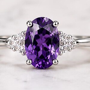 May include: A silver-toned ring featuring a large, oval-cut, deep purple amethyst gemstone held by four prongs. The band is accented on each side by a cluster of three small, clear, round-cut stones, creating a symmetrical design with a polished finish.