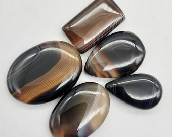 May include: Five polished agate stones in various shapes: oval, teardrop, and rectangular. The stones feature bands of brown, black, and white, with a glossy finish. These natural gemstones are ideal for jewelry making or collecting.
